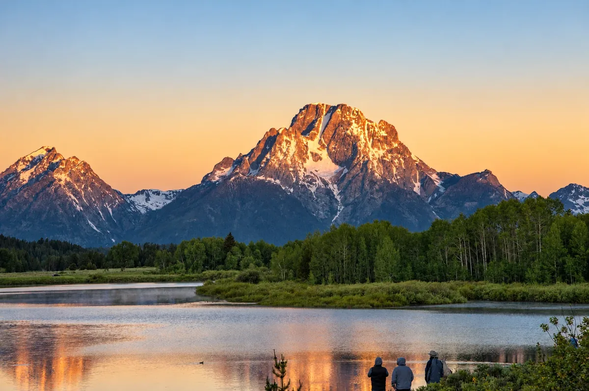 Snow-streaked mountain peaks glowing orange at sunset above a forest and a still river, three people watching from the bank